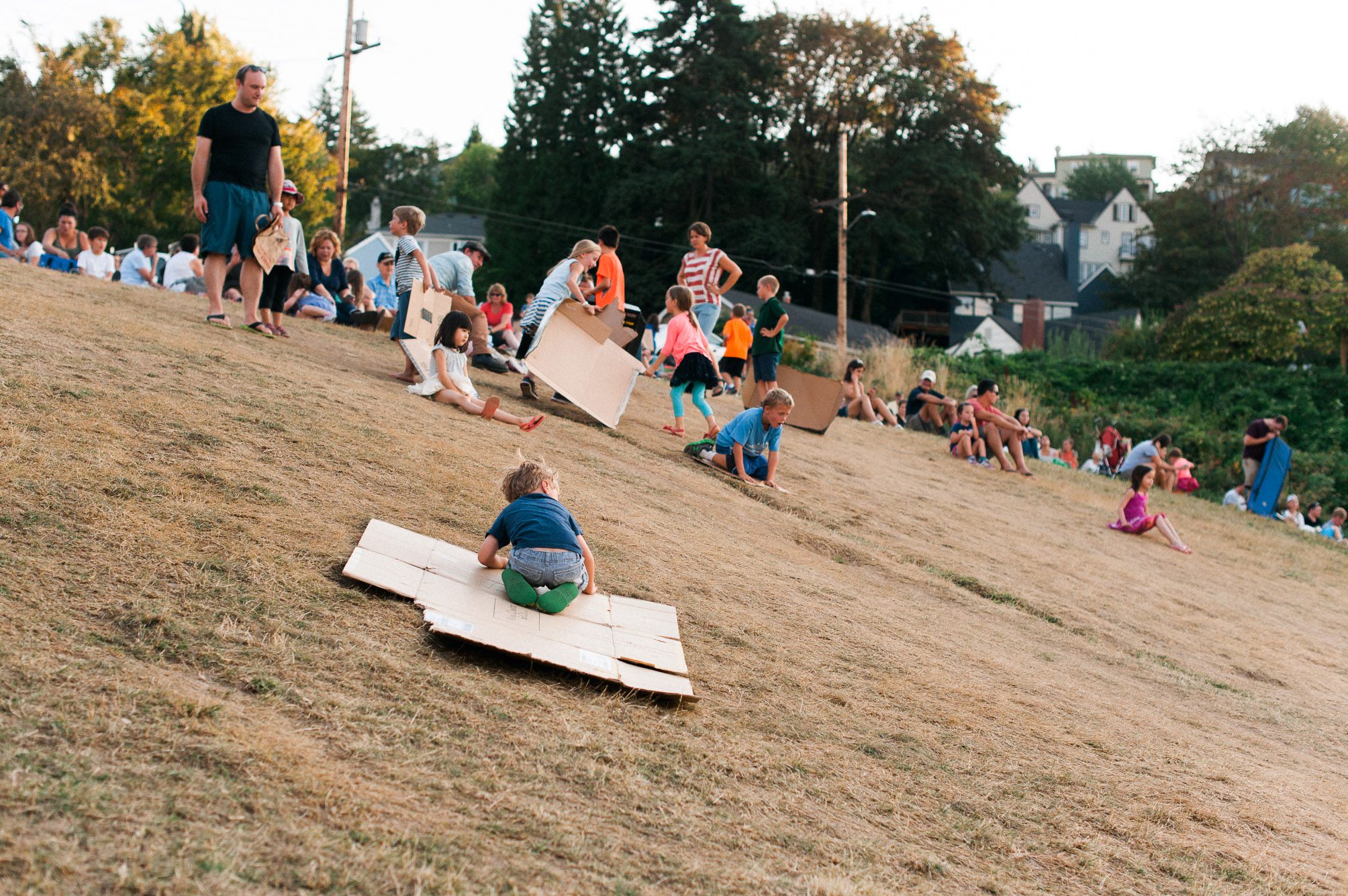 Local kids sliding down a grassy hill on pieces of cardboard while their parents wait for the Swifts to descend into Chapman Elementary School's Chimney. Photography by Briana Morrison