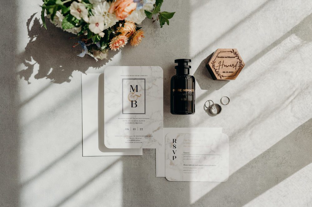 Sunshine and shadows highlight a beautiful neutral toned flatlay of flowers, wedding invitations, and wedding bands.