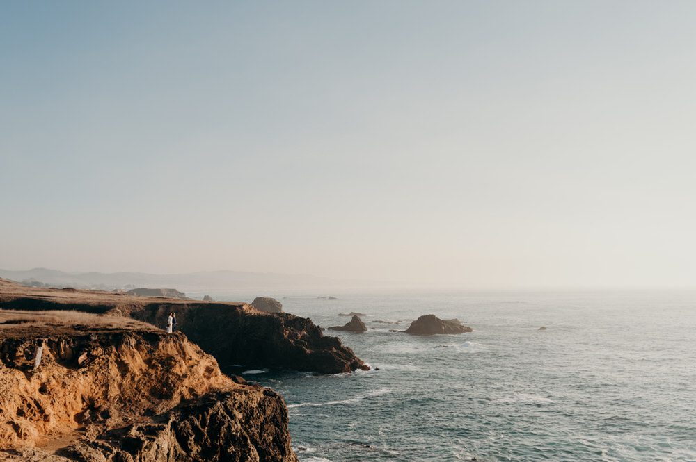 Oregon wedding photographer captures a beautiful coastal scene of cliffs, a slightly smoky sky, and the endless sea. A very tiny bride and groom stand on the cliff looking out to the ocean.
