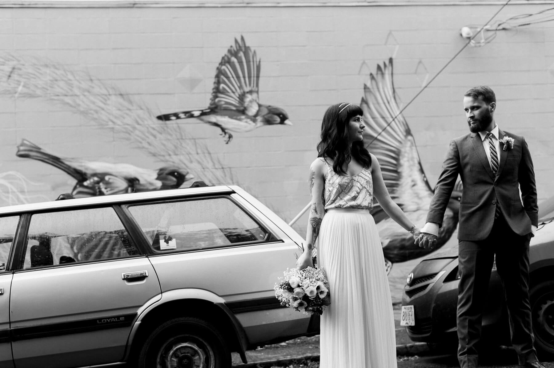 Portland, Oregon bride and groom pose in front of local cars and a bird mural in SE Portland. By Portland Holocene Photographer Briana Morrison