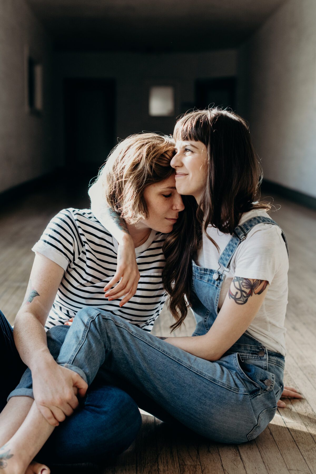 Cute cuddly same sex couple sits together during their engagement portraits.