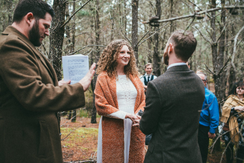 A bride (and wedding photographer) and groom tie the knot while holding a crystal in the woods.