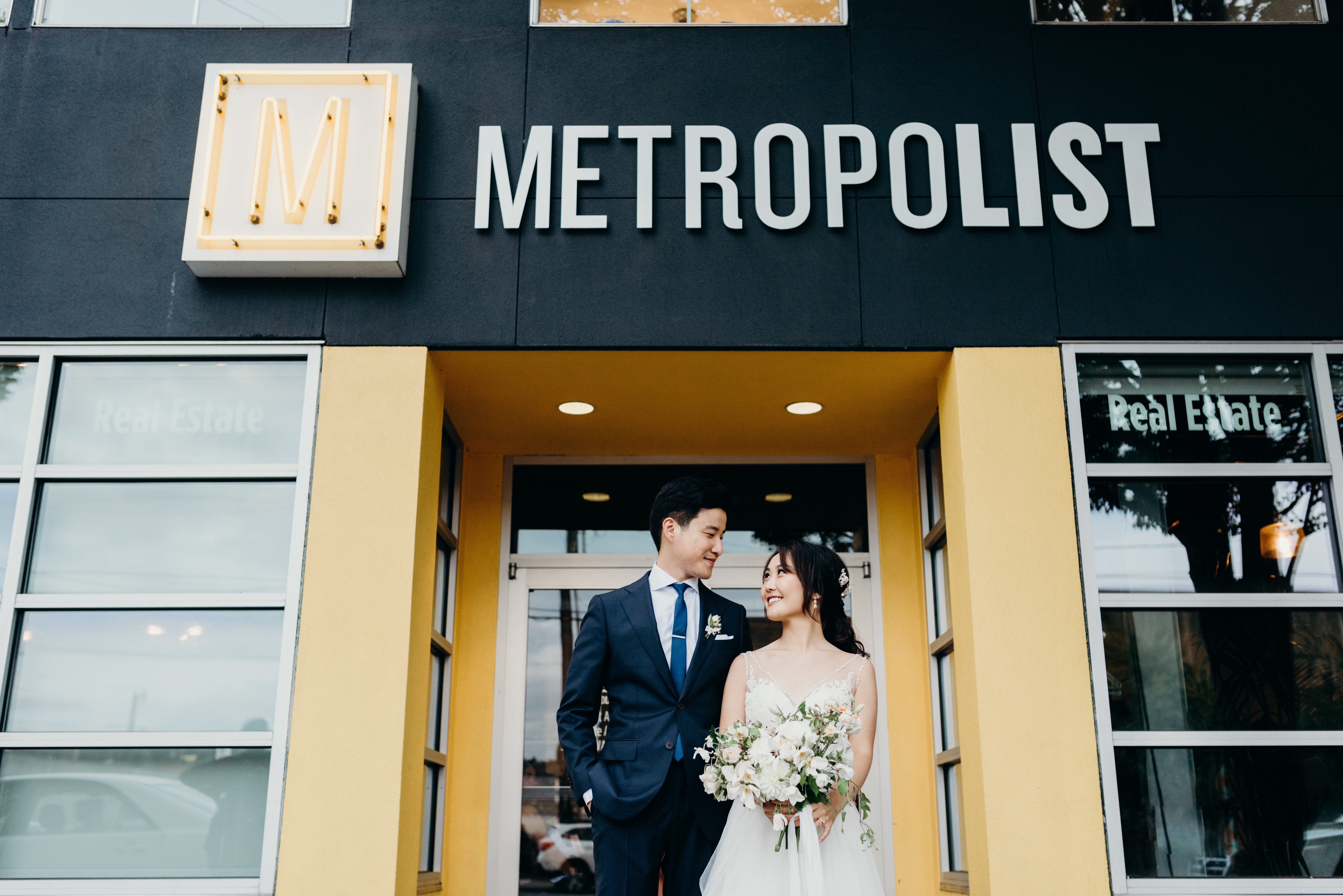 A bride and groom of Asian decent stand in front of the entry to their wedding venue, The Metropolist in Seattle, Washington