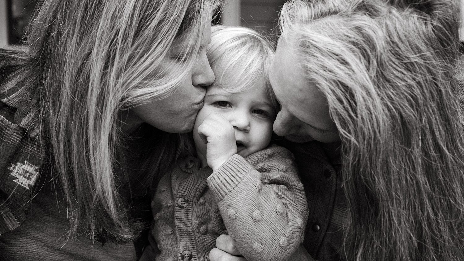 Portland family photographer captures black and white close up image of two moms kissing the cheeks of their toddler daughter who is looking into the camera with her thumb in her mouth.