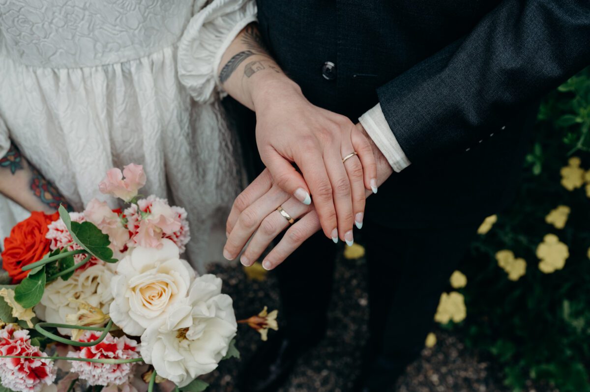 Portland wedding photographer captures a close up portrait of the hands of a bride and groom showing off their wedding rings with flowers surrounding them on both sides.