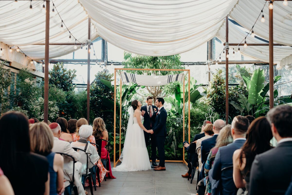 A large indoor wedding ceremony at Blockhouse in Portland, Oregon. The bride and groom stand under the chuppah holding hands and the bride is throwing her head back in laughter. They are surrounded by lush plants, cafe lights, and a large group of wedding gusts.