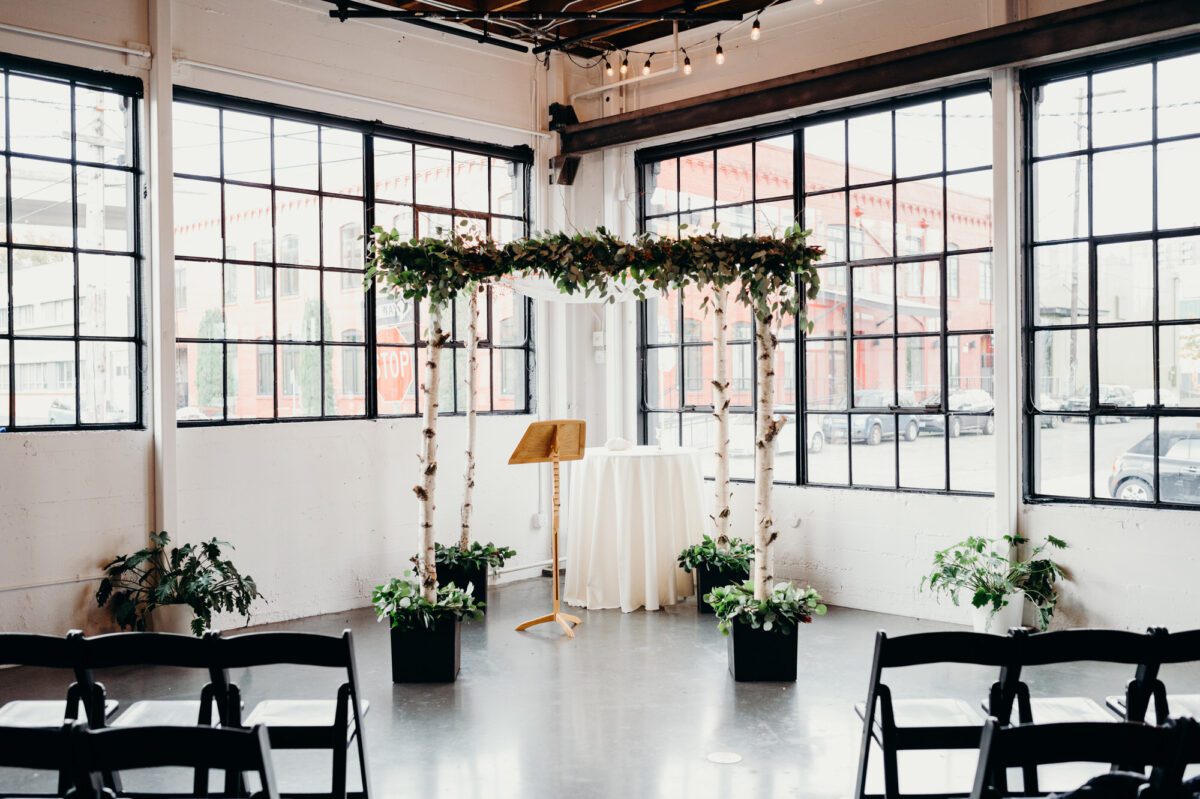 Castaway is a beautiful indoor wedding venue in Portland with large warehouse windows, painted white walls, and loads of natural light. Image shows a birch tree chuppah set up for a wedding ceremony in the corner of the venue with windows on both sides.