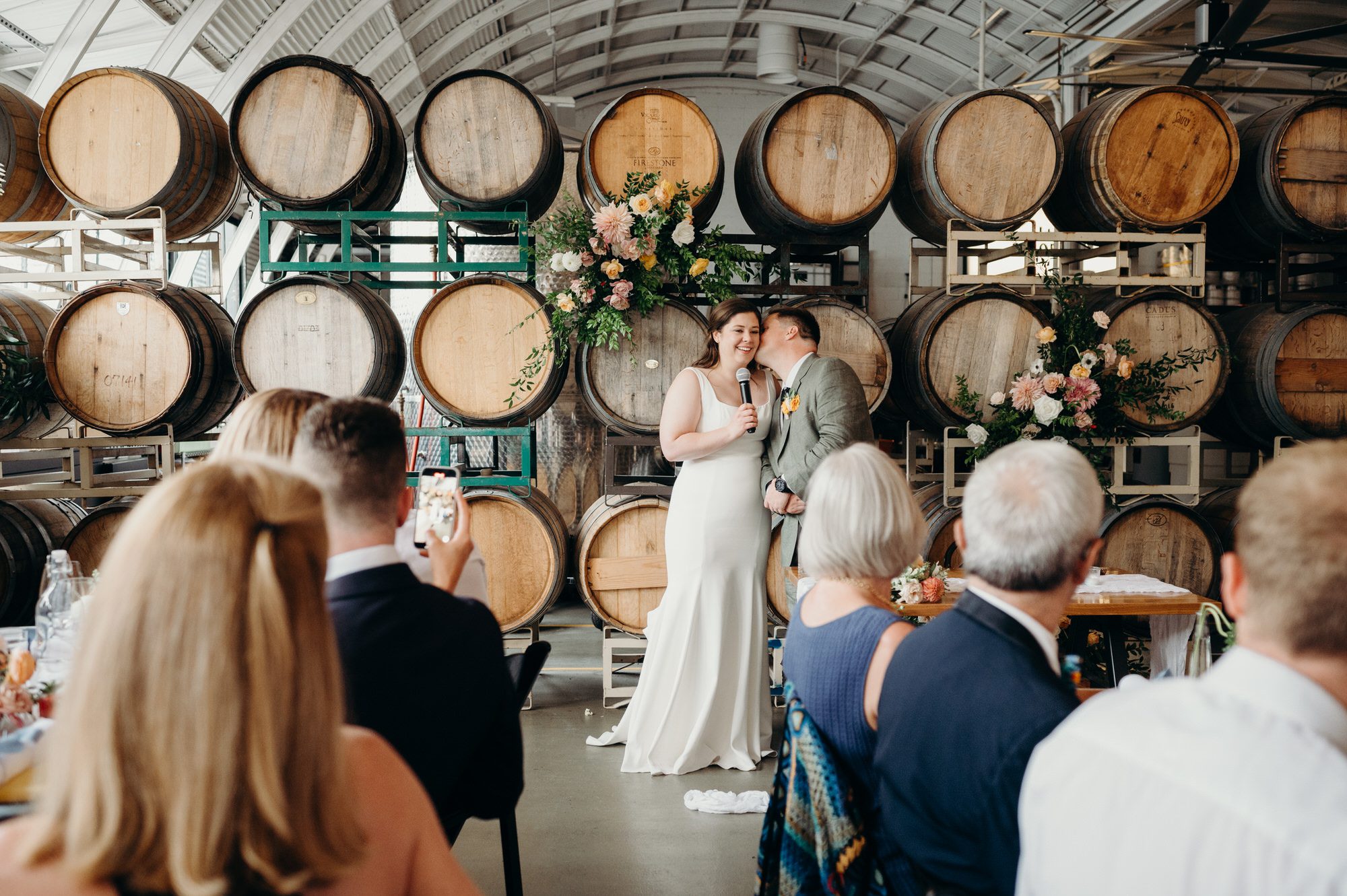 A bride and groom give a speech to their wedding guests at Coopers Hall - An indoor wedding venue in Portland with an arched metal ceiling and a wall of wood wine barrels behind them.