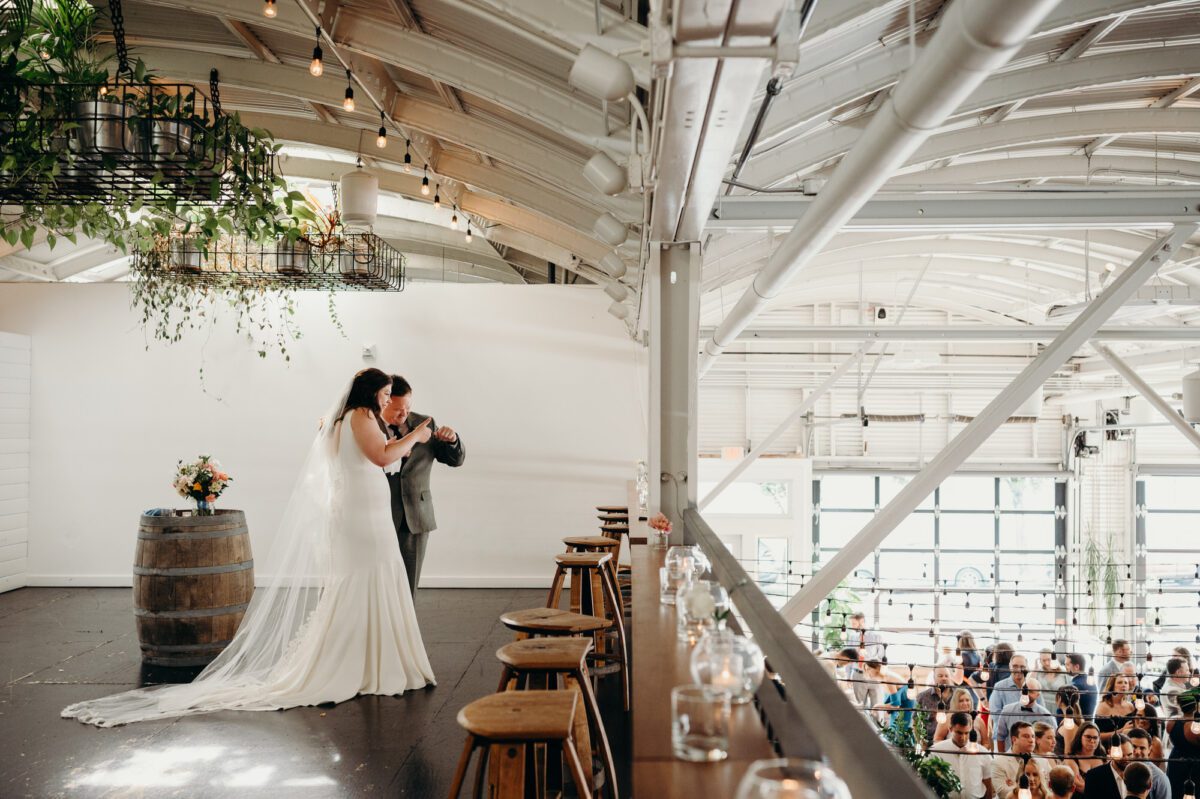 Bride and groom wave to their guests from the mezzanine in Coopers Hall. This indoor wedding venue in Portland has white walls, tall arched ceilings, and large windows.
