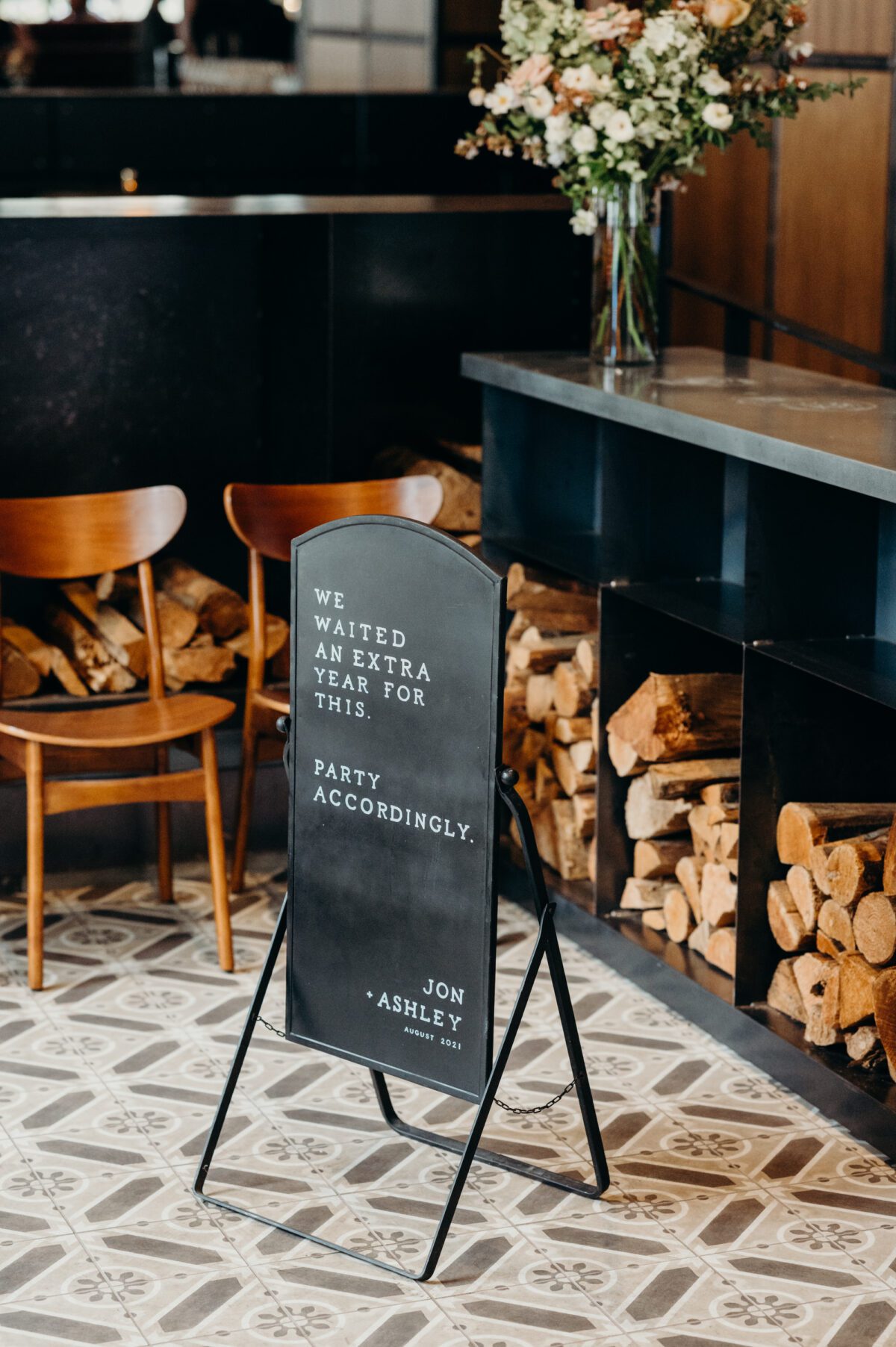 A modern black sign welcomes wedding guests to the indoor wedding venue Hunt & Gather Events. The background shows up a beautiful gray patterned tile floor, mid century wood chairs, and cubbies full of firewood.