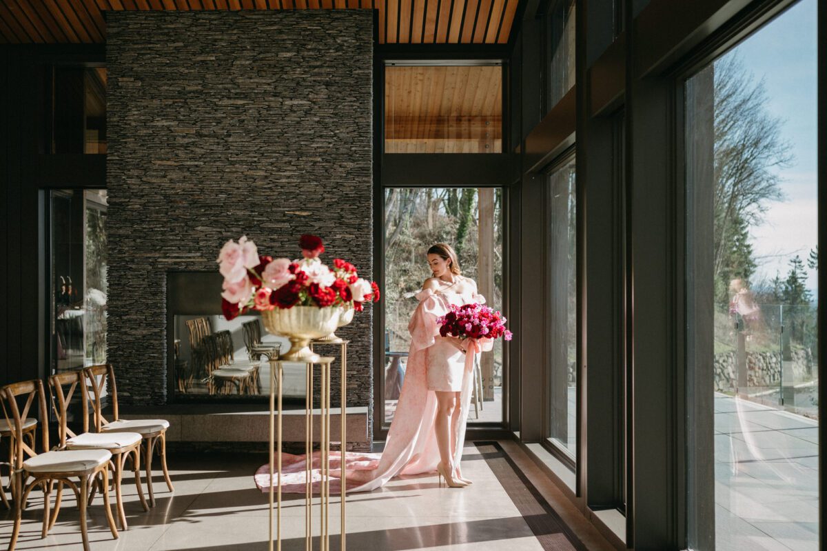 A bride in a modern pink wedding gown stands by the windows in the indoor ceremony space at Amaterra Winery. She holds a bouquet of red and pink flowers and the ceremony site is set with wood chairs and beautiful red and pink florals on stands.