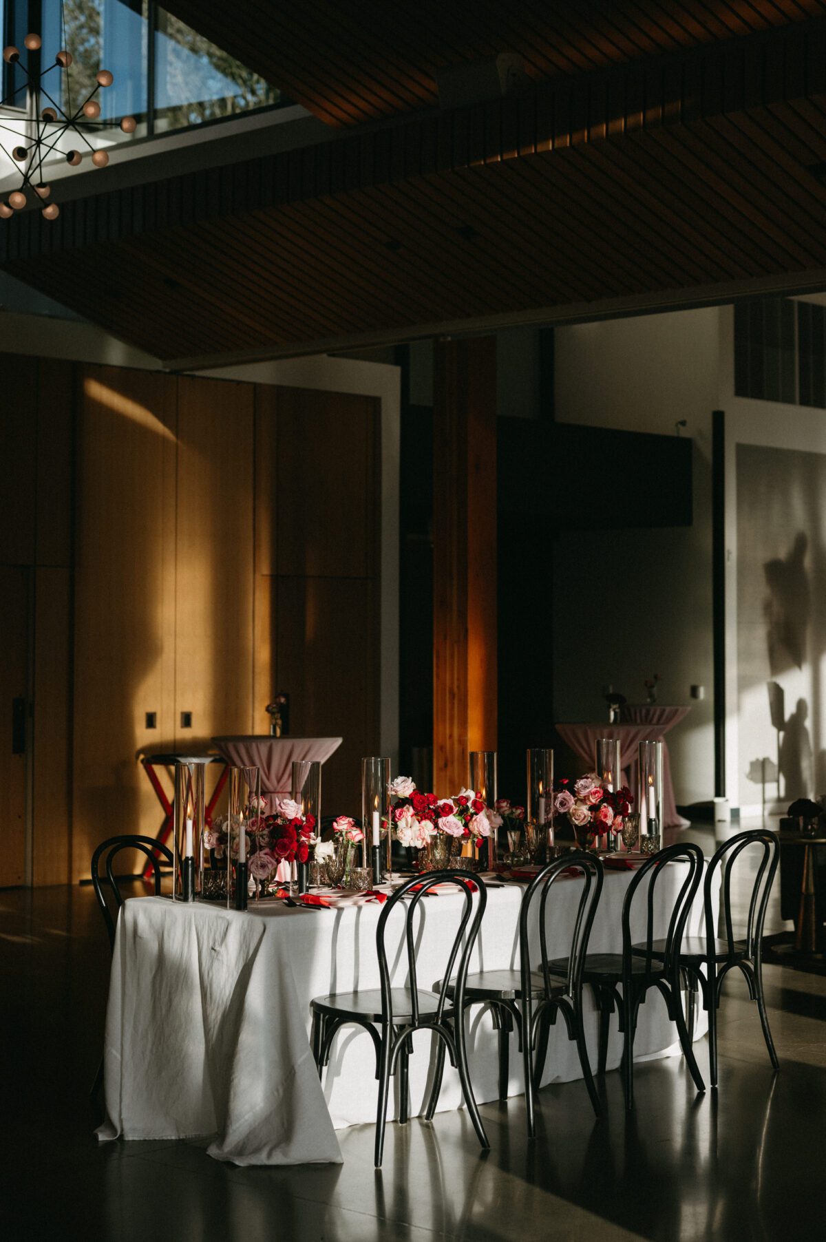 A dining table is glowing in the sunshine at the Amaterra Wines indoor wedding venue event space. The table is set with black bentwood dining chairs and red and pink florals.