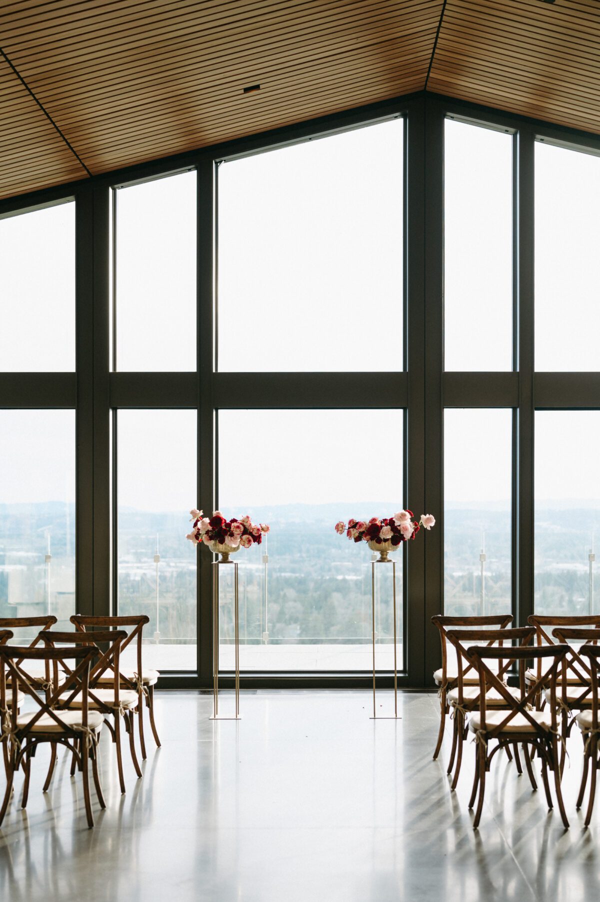 The Amaterra Wines indoor wedding venue Portland space with floor to ceiling windows overlooking the city of Portland, wood chairs set out for guests, and two stands flanking the ceremony site with large pink and red florals set on top.