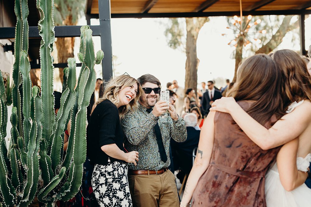 Two wedding guests take a photo of the bride and her friends on the wedding day. Everyone is smiling and there's a beautiful large cactus to the left of the group. Captured by Oregon wedding photographer Briana Morrison.