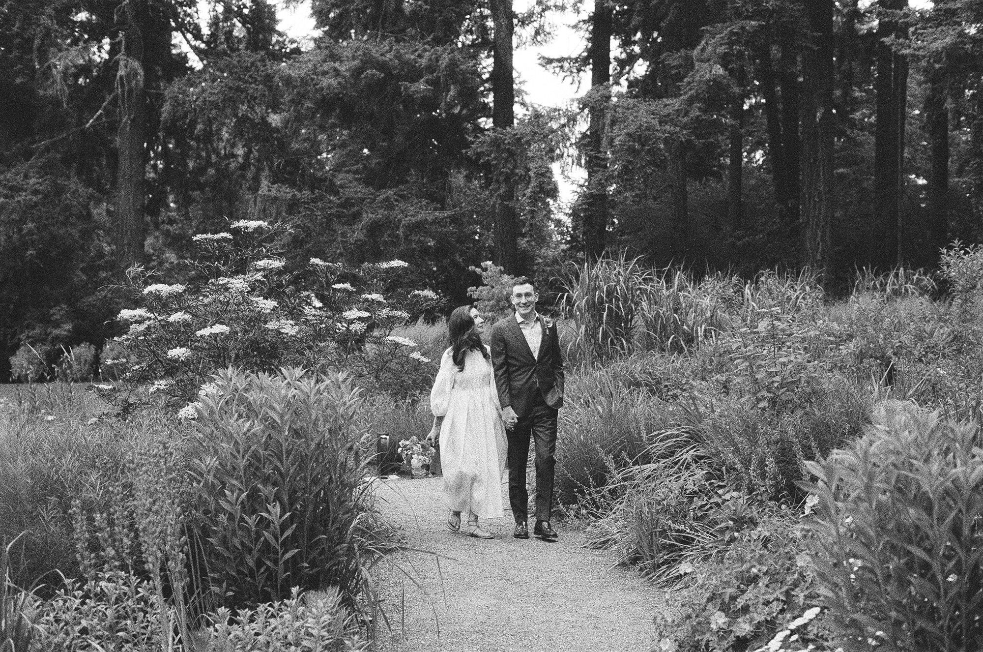 Portland wedding photographer captures a black and white film photograph of a bride and groom walking through Leach Botanical Garden