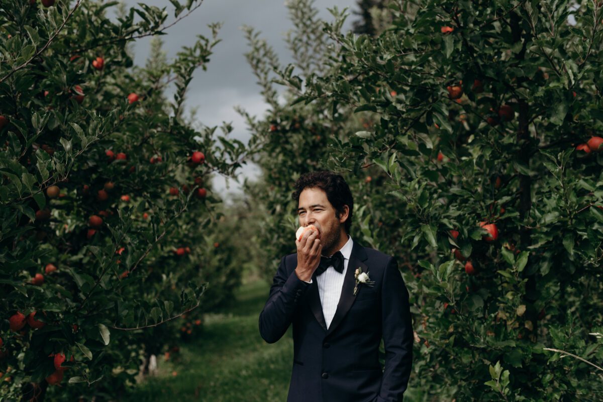 A man in a tuxedo bites into an apple while standing in an apple orchard.