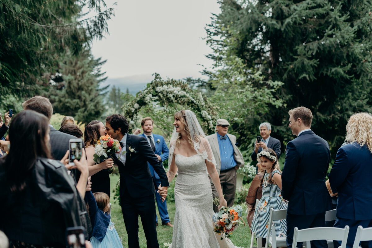 A bride and groom walk down the aisle after their ceremony at mt hood organic farms kissing their gusts on the cheek and smiling