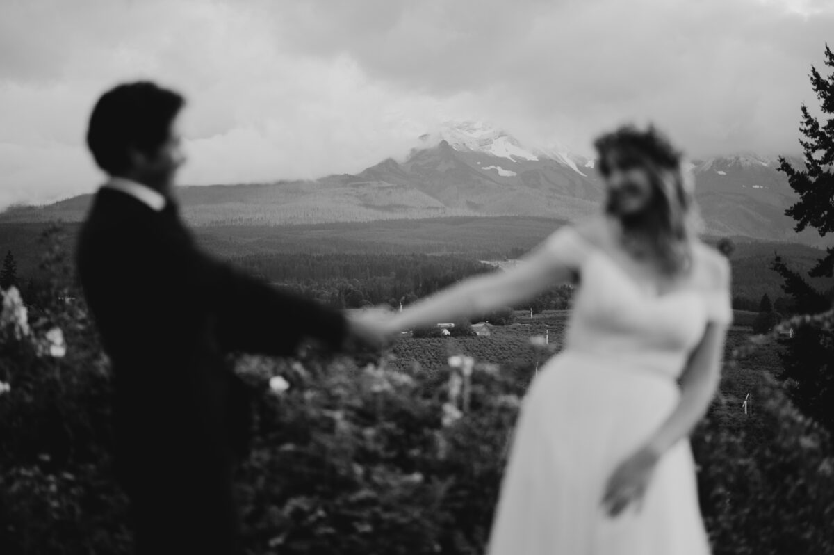 A blurry black and white portrait of a bride and groom holding hands and smiling at each other. The mountain in the background is in focus and slightly covered in clouds at mt hood organic farms