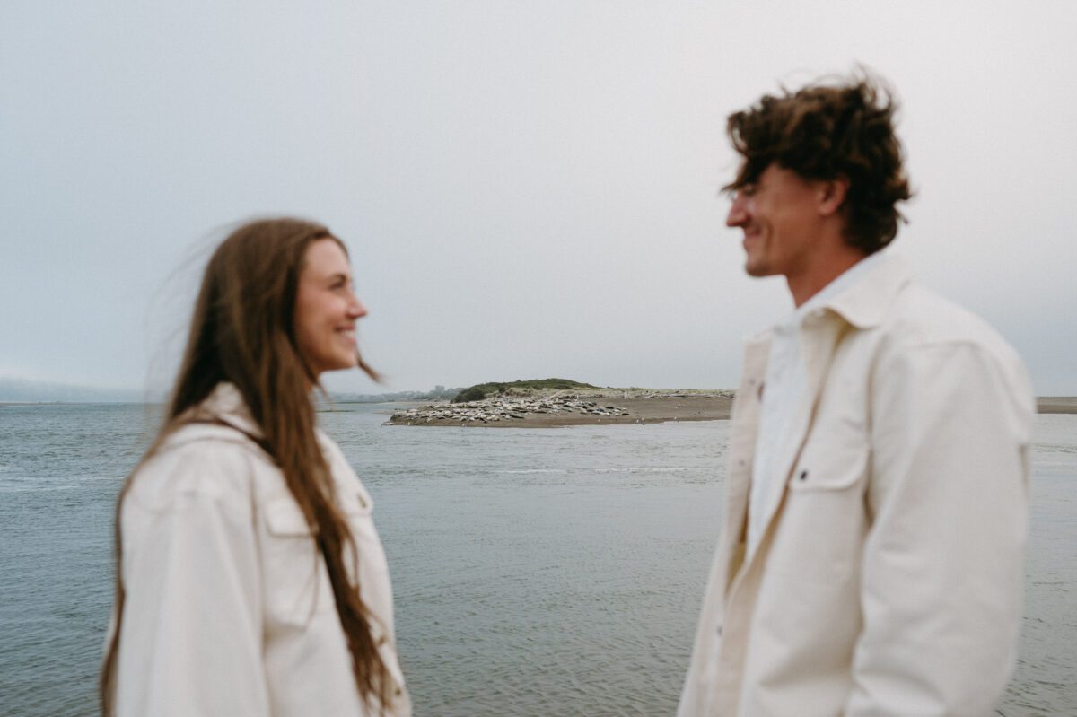 A bride and groom look at each other and smile - the camera is focused on a beach full of sea lions in the background.