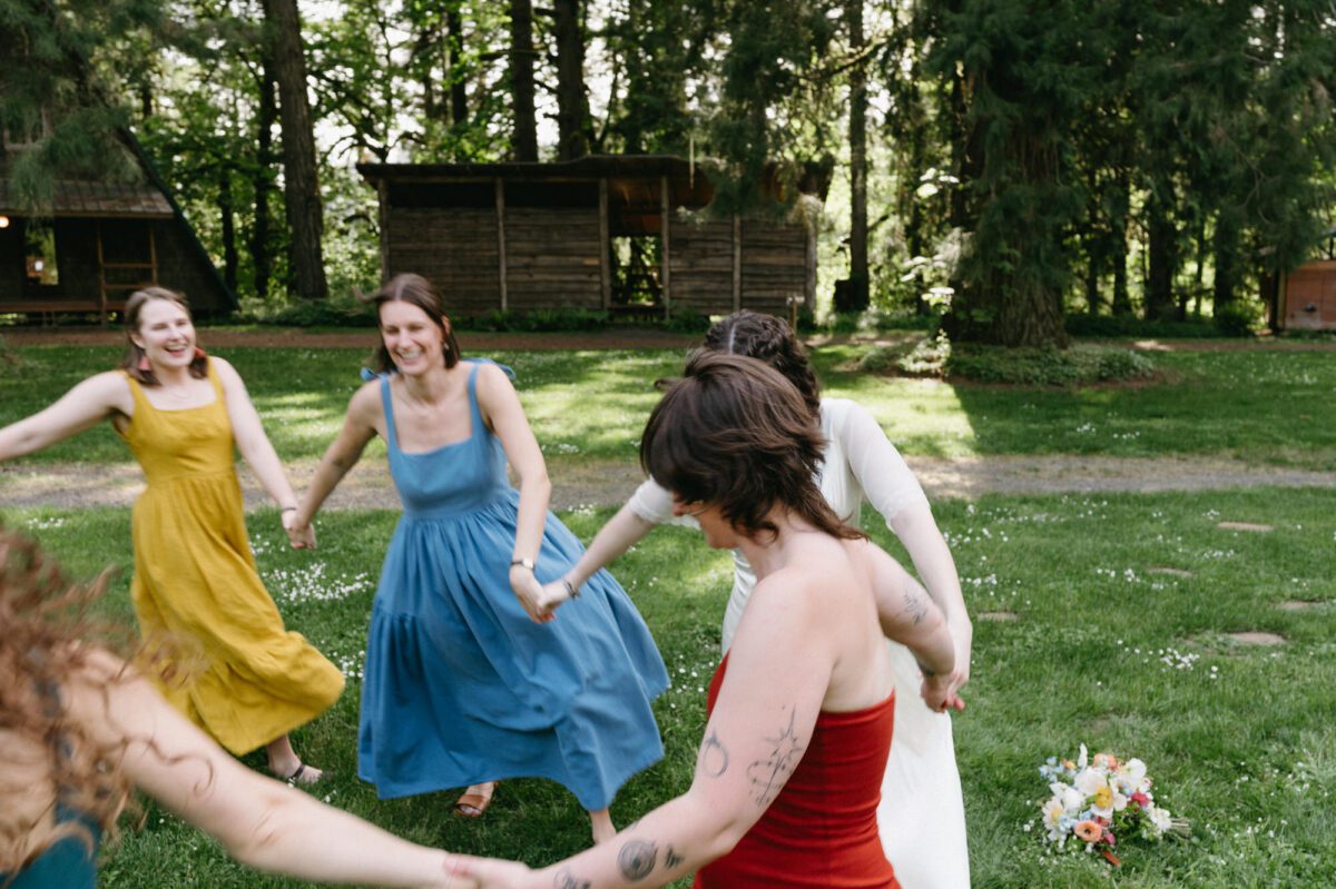 A rainbow of dresses in this motion blur photograph of a bride dancing in a circle with her wedding party. Documentary summer camp wedding photography by Briana Morrison