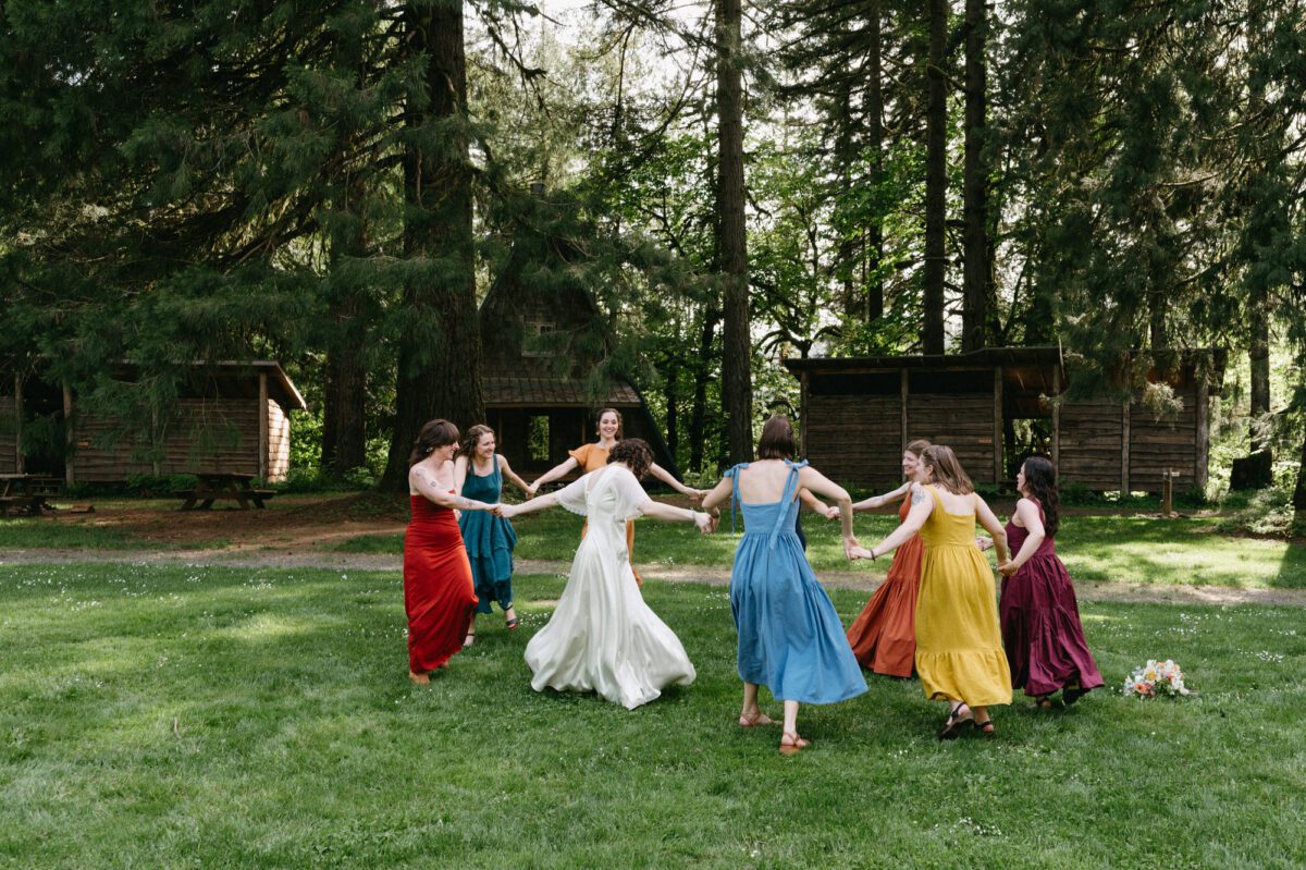 A bride dances in a circle with her wedding party creating a rainbow of color in a summer camp meadow. Documentary summer camp wedding photography by Briana Morrison