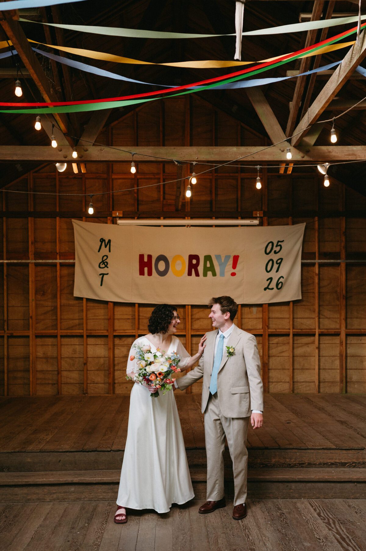 A bride and groom look at each other and laugh while standing in front of their handmade wedding banner. Summer camp wedding photography by Briana Morrison